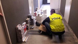 Policjant w kamizelce z napisem Policja kuca na korytarzu, obok paczki papierosów.