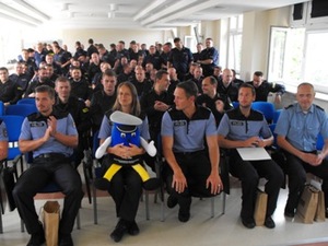 Zdjęcie wspólne uczestników imprezy siedzących na sali szkoleniowej (uczestniczka siedząca na środku w pierwszym rzędzie trzyma na kolanach maskotkę Komendy Wojewódzkiej Policji w Łodzi - Komisarza Błyska)