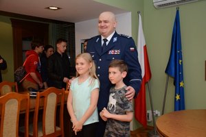 Gabinet komendanta. Inspektor Andrzej Łapiński pozuje do zdjęcia stojąc z dwójką dzieci na tle flag polskiej i unijnej.