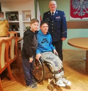 Gabinet komendanta. Inspektor Andrzej Łapiński stojąc pozuje do zdjęcia z dwoma chłopcami. Nastolatek siedzi na wózku inwalidzkim przed komendantem. Młodszy chłopiec stoi obok niego.