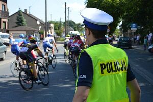 policjant w białej czapce i kamizelce z napisem policja stoi tyłem i obserwuje jadący peleton kolarzy.