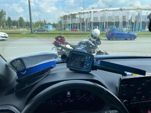 Widok z wnętrza radiowozu, na podszybiu leży czapka policyjna oraz ręczny miernik prędkości, na zewnątrz widać motocykl oraz auto.
