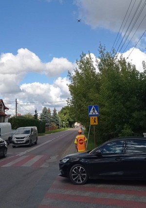 widok na drogę, przy przejściu dla pieszych stoi pan &quot;stopek&quot;, nad drogą lata policyjny dron