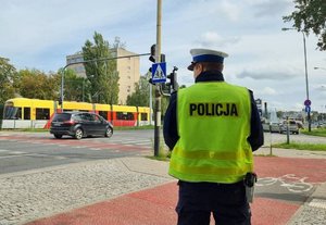 Policjant w mundurze oraz kamizelce odblaskowej z napisem Policja stoi przy drodze dla rowerów. W tle auto oraz tramwaj.