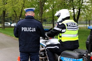 na zdjęciu policjant stoi tyłem, obok funkcjonariusza siedzącego na służbowym motocyklu.
