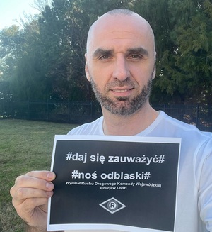 Mężczyzna trzyma kartkę z czarnym tłem na niej napis # daj się zauważyć # noś odblaski.