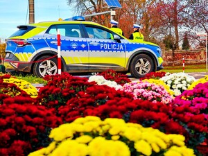 Policyjny radiowóz, za nim umundurowani funkcjonariusze. Na pierwszym planie kolorowe kwiaty.