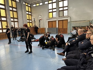 Uczniowie klasy mundurowej, w czarnych spodniach i czarnych koszulkach, podczas pokazu na sali gimnastycznej. Dwie dziewczyny trzymają długą broń w rękach.