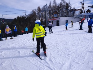 Dzieci w kamizelkach odblaskowych i kaskach zjeżdżają ze stoku na nartach. W tle wyciąg narciarski i drzewa.