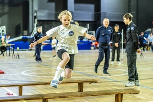Zmagania dzieci podczas zawodów sportowych na sali gimnastycznej w obecności policjantów i aktorów.