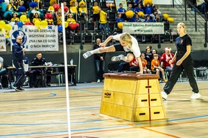 Zmagania dzieci podczas zawodów sportowych na sali gimnastycznej w obecności policjantów i aktorów.