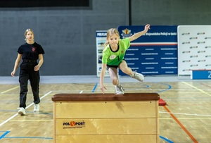 Zmagania dzieci podczas zawodów sportowych na sali gimnastycznej w obecności policjantów i aktorów.