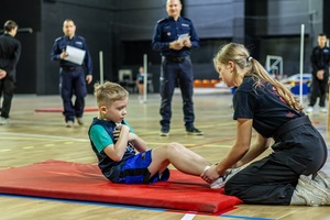 Zmagania dzieci podczas zawodów sportowych na sali gimnastycznej w obecności policjantów i aktorów.