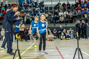 Zmagania dzieci podczas zawodów sportowych na sali gimnastycznej w obecności policjantów i aktorów.