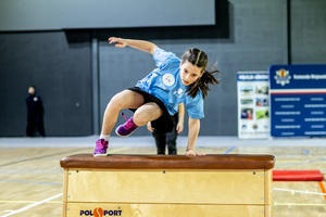 Zmagania dzieci podczas zawodów sportowych na sali gimnastycznej w obecności policjantów i aktorów.