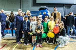 Zmagania dzieci podczas zawodów sportowych na sali gimnastycznej w obecności policjantów i aktorów.