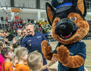 Maskotka policyjna - Komisarz Alex, pies wita się z dziećmi. Na drugim planie umundurowany policjant podaje dłoń dzieciom na przywitanie.