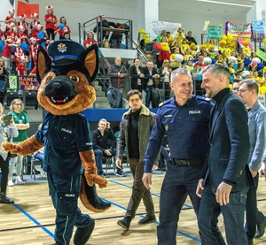 Umundurowany policjant rozmawia z mężczyzną, aktorem. Obok idzie policyjna maskotka - pies policjant.