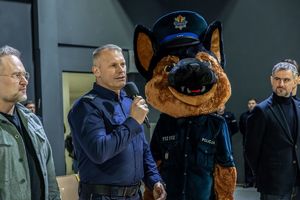 Umundurowany policjant z mikrofonem stoi obok maskotki policyjnej przedstawiającej psa policjanta.