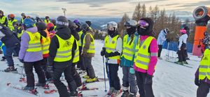 Grupa dzieci stoi na stoku, ubrani w kamizelki odblaskowe.