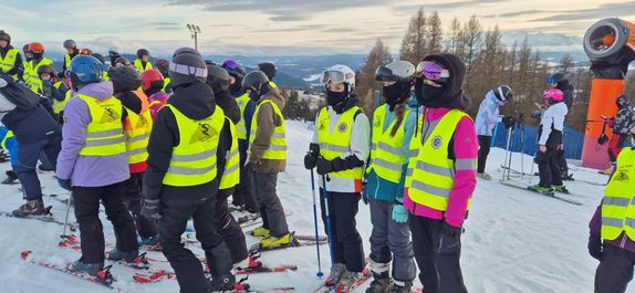 Grupa dzieci stoi na stoku, ubrani w kamizelki odblaskowe.