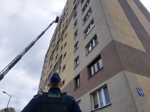 policjant stojący na dole budynku patrzący w górę jak strażacy na drabinie podjeżdżają do najwyższego piętra, widać z okien wydobywający się dym