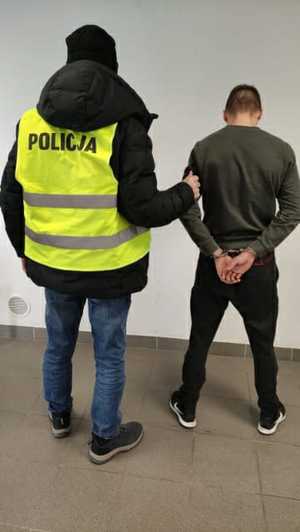 policjant stojący na korytarzu ze sprawcą rozboju, który ma założone kajdanki na ręce trzymane z tyłu