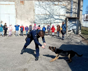 policjant-przewodnik psa służbowego ze swoim podopiecznym, w tle dzieci z grupu przedszkolnej