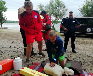policjant oraz ratownik wodny uczy chłopca jak prawidłowo przeprowadzić resuscytację krążeniowo - oddechową.