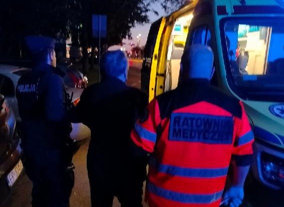 Policjant wraz z ratownikiem prowadzi seniora do karetki.