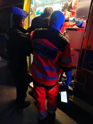 Policjant wraz z ratownikiem prowadzi seniora do karetki.