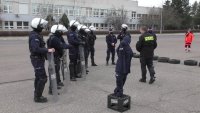 policjanci stojący na betonowym placu ubrani w stroje ochrony indywidualnej z kaskami na głowie przysłuchują się słowom prowadzącego zajęcia instruktora. na pierwszym planie manekin ubrany w policyjny mundur ćwiczebny