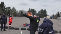 Instruktor z płonącym przedramieniem stojący bokiem do obiektywu aparatu. W tle asekurujący ratownik medyczny i policjant.