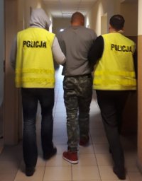 Policjanci ubrani w żółte kamizelki odblaskowe prowadzą pod ręce zatrzymanego mężczyznę ubranego w szarą bluzę i spodnie typu bojówki