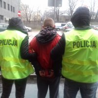 Policjanci w kamizelkach odblaskowych z napisem Policja wraz z zatrzymanym, który ma założone kajdanki na ręce trzymane z tyłu