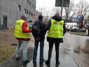 policjanci w kamizelkach odblaskowych z napisem policja prowadza na dziedzińcu komisariatu zatrzymanegp