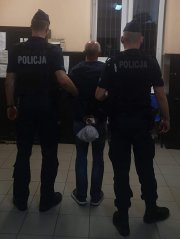 poszukiwany mężczyzna na korytarzu komisariatu jest trzymany przez dwóch policjantów z Oddziału Prewencji