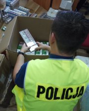 Nieumundurowany policjant w kamizelce z napisem POLICJA przegląda zabezpieczone papierosy w dużym kartonowym pudle. W pokoju na podłodze i biurku leżą pozostałe paczki nielegalnych  papierosów.
