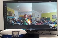Monitor komputera na którym widać dzieci w klasie, które siedzą w ławkach i uczestniczą w lekcji on-line. Chłopiec w zielonej koszulce stoi przy ławce. Przed monitorem leży czapka policjanta ruchu drogowego