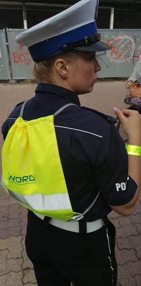 Na zdjęciu widzimy policjantkę z ruchu drogowego, która ubrana jest w mundur policyjny oraz w białą czapkę policyjną , na plecach ma założony worek odblaskowy.