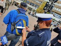 Na zdjęciu policjantka Wydziału Ruchu Drogowego KMP w Łodzi przypina element odblaskowy do plecaka mężczyzny. W tle widać fragment ulicy Piotrkowskiej.