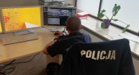 Policjant w koszuli z krótkim rękawem siedzi przy biurku, na którym znajdują się monitory z obrazem kilku ulic i skrzyżowań.