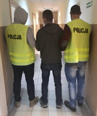 Nieumundurowani policjanci w kamizelkach z napisem POLICJA prowadzą zatrzymanego sprawcę rozboju