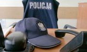 Na obrazku widzimy położoną na biurku granatową czapkę policyjną, koszulkę z napisem policja , która jest powieszona na krześle . Na biurku leży również telefon z odłożona obok słuchawką.