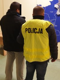 Funkcjonariusz w kamizelce z napisem POLICJA prowadzi zatrzymanego sprawcę kradzieży