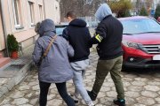 Zdjęcie jest zrobione przed wejściem do II Komisariatu Policji. Widać na nim zatrzymanego ubranego w szare spodnie dresowe oraz ciemną kurtkę, który jest prowadzony przez policjanta i policjantkę . Obydwoje mają założone kaptury na głowie. W tle widać czerwony samochód.