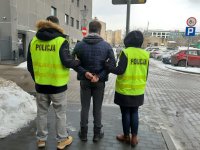 Na dziedzińcu Komendy Miejskiej Policji w Łodzi, policjanci w zielonych kamizelkach policyjnych prowadzą zatrzymanego ubranego w szare spodnie dresowe oraz ciemną kurtkę, na rękach ma założone kajdanki.
