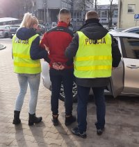 Zatrzymany jest prowadzony przez policjantkę i policjanta do samochodu, ubrani są w policyjne kamizelki z napisem policja, mężczyzna ma założone z tyłu kajdanki i ubrany jest w  czerwoną bluzę polarową.