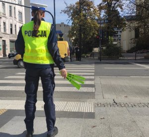 Umundurowana policjantka ruchu drogowego na skrzyżowaniu stojąca tyłem do kadru, ubrana w kamizelkę odblaskową z napisem Policja. Policjantka trzyma w ręce odblaski.