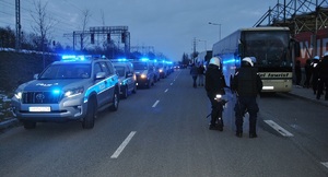 Wzdłuż jezdni po jednej stronie stoją radiowozy z włączonymi światłami błyskowymi, po prawej policjanci przy autokarze gości.